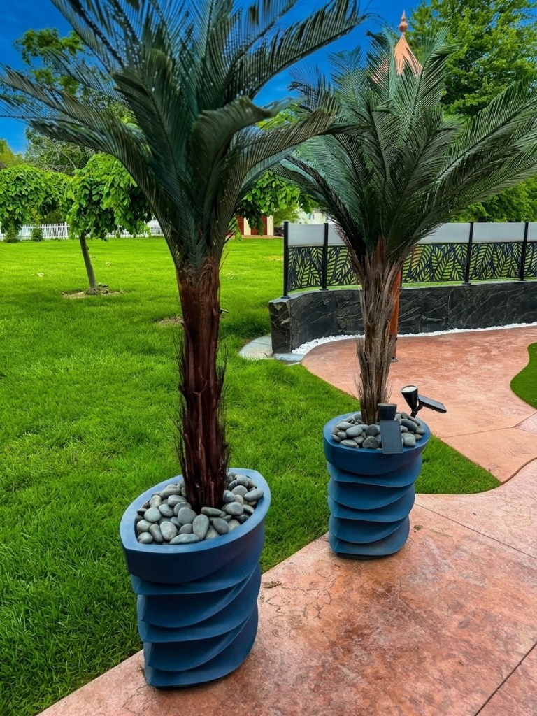 Finished artificial cycas palm with river rocks on top of foam and brick base in tall blue planter on stamped concrete patio