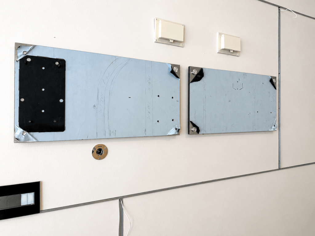 Two wall-mounted metal plates with screw holes, one with a black attachment, against a light-colored wall.