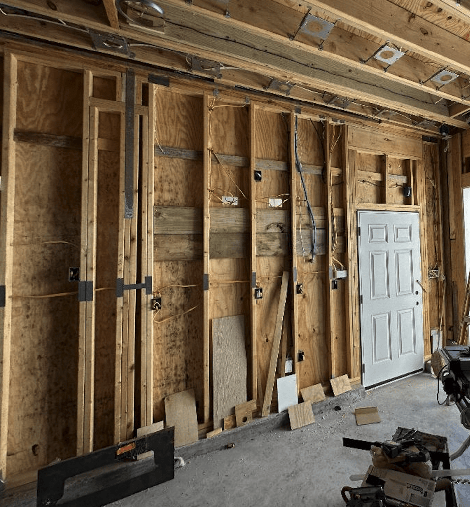 An interior wall frame of a room under construction, showing exposed wooden beams, electrical wiring, and a partially installed door.