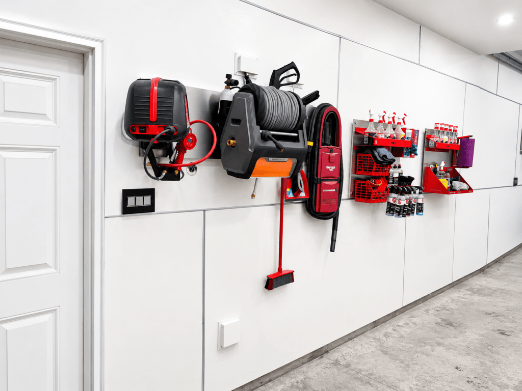 A well-organized wall in a garage featuring a variety of tools and cleaning supplies mounted on red and gray shelving. Items include a hose reel, vacuum cleaner, spray bottles, and cleaning cloths. A white door is also visible on the left.